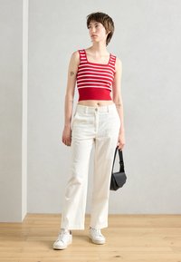 Top court rouge et blanc rayé, pantalon large en velours cotelé blanc, baskets blanches, tenant un petit sac noir, debout sur un sol en bois.