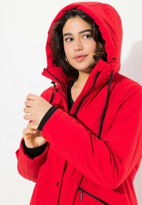 Red waterproof jacket with a high collar and a large, adjustable hood. Features a zipper, drawstrings, and black accents.