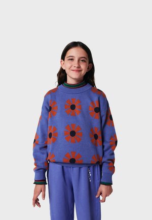 Purple sweater with bold orange flower patterns and black centers, featuring a loose fit and ribbed cuffs. Paired with purple pants.
