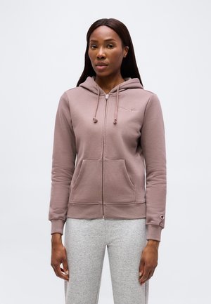 Kvinde iført en lys mauve lynlås-hættetrøje med forlommer og grå joggingbukser, stående mod en ensfarvet lys baggrund.