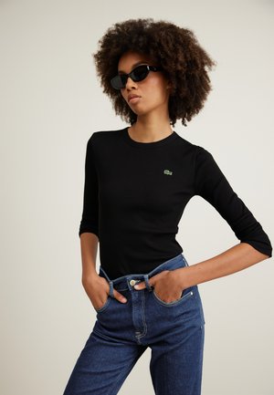 Black ribbed long-sleeve top with round neckline and green logo. Paired with blue denim high-waisted jeans featuring button and pockets.