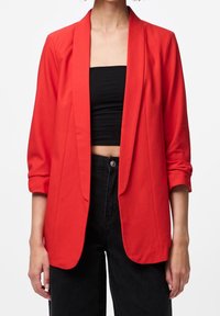 Blazer rouge en tissu léger, coupe ajustée, devant ouvert et manches retroussées. Porté sur un haut court noir et un jean taille haute.