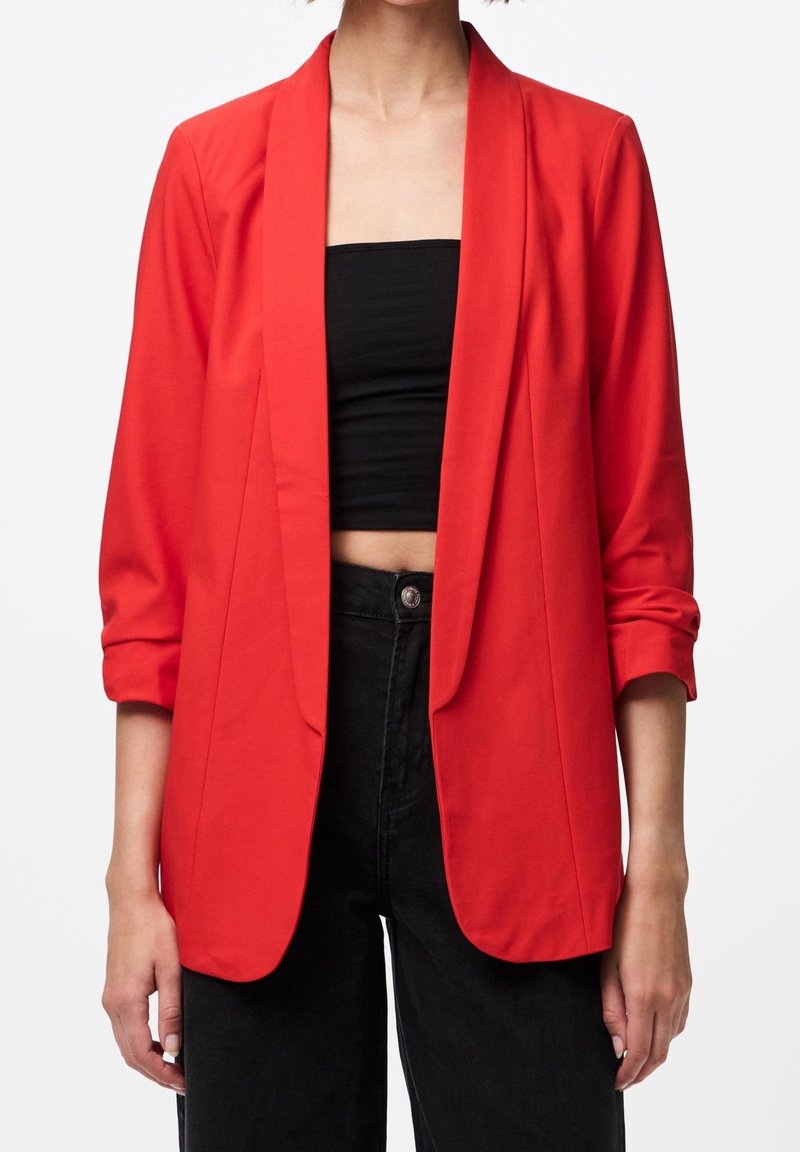 Blazer rouge en tissu léger, coupe ajustée, devant ouvert et manches retroussées. Porté sur un haut court noir et un jean taille haute.