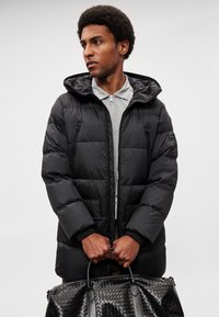 Casaco preto acolchoado com capuz, com fecho de correr frontal, múltiplos bolsos e textura acolchoada. Modelo segura uma bolsa preta texturizada.