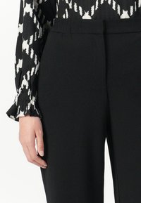 Persoon in zwarte broek met elastische tailleband en een zwart-wit gestreept lange mouw top, hand langs het lichaam.