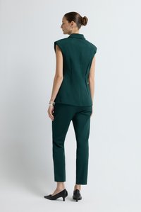 Žena s vlasy v drdolu, oblečená v bezrukávé tmavě zelené přizpůsobené vestě a odpovídajících kalhotách, stojící zády k fotoaparátu, na jednobarevném světlém pozadí.