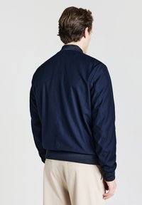 Un homme de dos portant une veste bomber bleu marine et un pantalon beige sur un fond clair uni.
