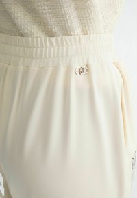 Pantalon crème clair avec une texture douce et lisse. Il dispose d'une taille élastique et d'un accent en métal argenté sur le côté.