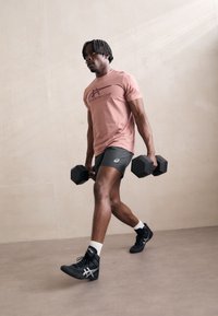 Sporty mand, iført en pink t-shirt og mørke shorts, bærende på sorte hexagonale håndvægte, med sorte sneakers og hvide ankelsokker.