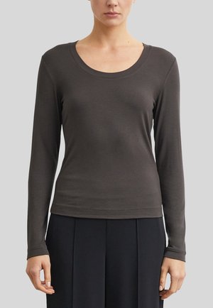 Femme portant un haut à manches longues gris foncé uni et un pantalon noir, debout devant un fond gris clair.