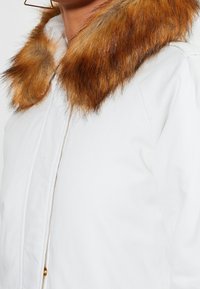 Biała kurtka z brązowym sztucznym kołnierzem z futra. Posiada zapięcie na guziki i gładką teksturę. Charakterystyczny jest kontrast kolorów brązu i bieli.