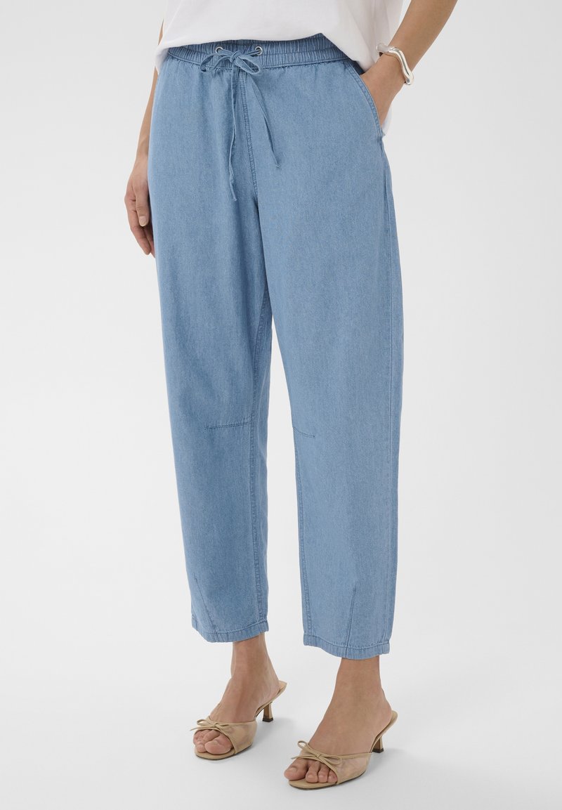 Femme portant un pantalon jogger en denim bleu clair avec taille à cordon de serrage et des sandales à talons beiges, sur un fond blanc.