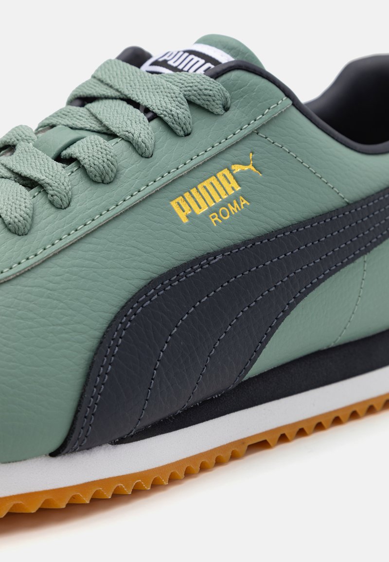 Hellgrüner Sportschuh aus Leder mit schwarzem Streifen-Design, goldenem Logo, strukturierten Schnürsenkeln und einer Gummisohle mit gezacktem Rand.
