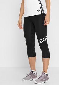 Czarne legginsy do kostek z białym napisem "BORG" z boku, z fakturowanym wykończeniem i gładkim dopasowaniem, w zestawieniu z szarymi butami sportowymi.