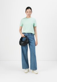 Ung kvinne står iført mintgrønn t-skjorte, vide blå jeans, hvite sneakers, og holder en liten svart håndvott mot hvit bakgrunn.