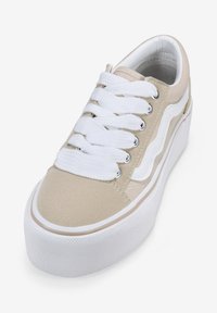 Zapatillas de lona beige con cordones blancos, ojales circulares y suela de goma blanca. Presenta un diseño de rayo lateral. Parte superior de tela texturizada.