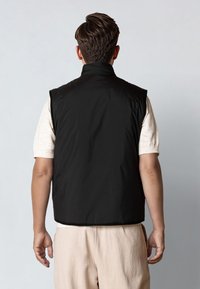 Hombre con cabello corto y castaño, vistiendo un chaleco negro sin mangas sobre una camisa de manga corta beige claro y pantalones beige, de espaldas a la cámara.