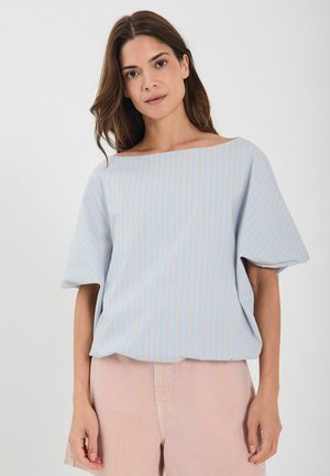 Hellblaues Oberteil mit vertikalen Streifen in Orange und Weiß. Weit geschnittene Ausschnitt, lockere Passform und kurze Ärmel. Kombiniert mit hellrosa Shorts.