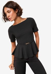 Czarna bluzka peplum z krótkimi rękawami i półstrukturalnym krojem, podkreślona złotym paskiem z klamrą, noszona z czarnymi spodniami.