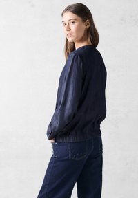 Marinefarbener, leichter Mantel mit lockerer Passform, gerafftem Saum und langen Ärmeln. Kombiniert mit dunklen Denim-Hosen mit subtilen Nähten.