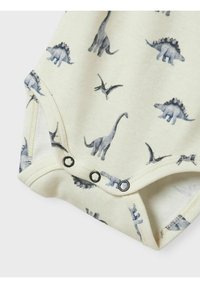 Bebi bodi s kratkim rukavima u krem boji, s plavim otiscima dinosaura. Tri crna dugmeta na dnu za jednostavno kopčanje.