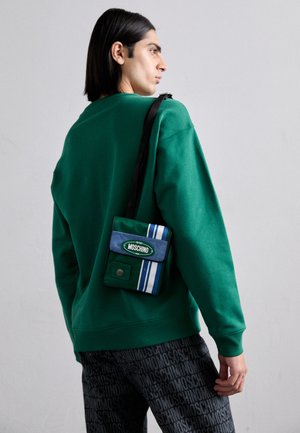 Grüner Pullover, gemusterte schwarze Hose und eine kleine blaue Tasche mit grünen und weißen Streifen, die ein Logo auf der Vorderseite hat.