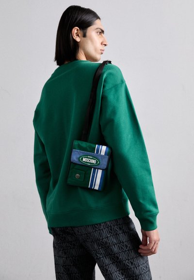 Pulover verde, pantaloni negri cu un model și o geantă mică albastră cu dungi verzi și albe, având un logo pe partea din față.