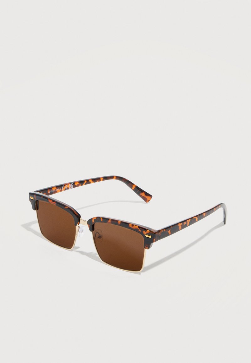 Τετράγωνα γυαλιά ηλίου τύπου χελώνας με καφέ φακούς και λεπτά ακρυλικά στοιχεία σε χρυσό μεταλλικό πλαίσιο, τοποθετημένα σε λευκή επιφάνεια.