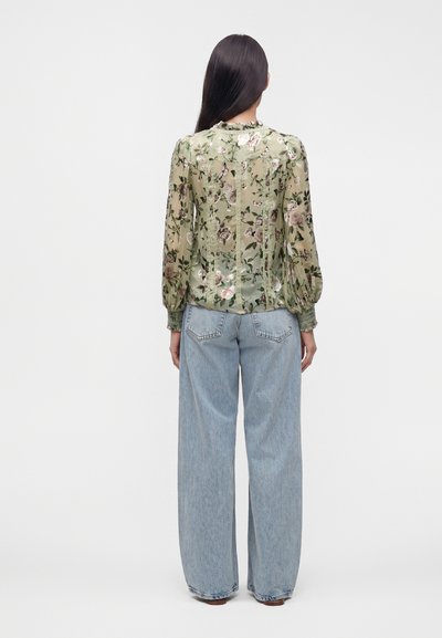 Femme aux longs cheveux foncés portant un chemisier vert transparent à fleurs et un jean large bleu clair, debout de dos sur un fond uni.