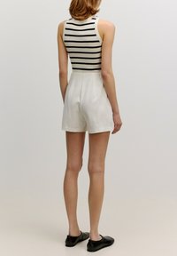 Schwarz-weiß gestreiftes ärmelloses Oberteil mit hohem Kragen, kombiniert mit cremefarbenen Shorts. Model trägt schwarze flache Schuhe.