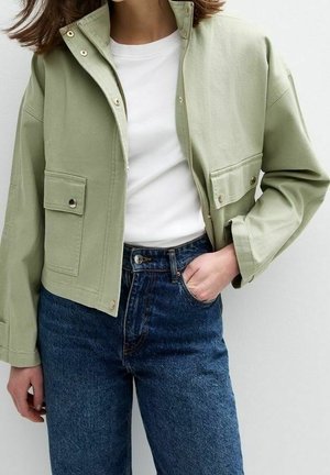 Femme portant une veste courte vert clair avec des poches avant, par-dessus un t-shirt blanc et un jean bleu taille haute, une main dans la poche.
