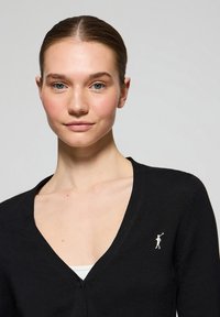 Cardigan noir en V en tissu tricoté, avec une broderie discrète du logo sur la poitrine gauche et une fermeture à boutons à l'avant.