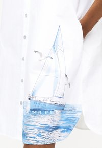 Λευκό πουκάμισο με εκτύπωση μπλε ιστιοφόρου και γλάρων κοντά στο τελείωμα, που δείχνει μέρος του χεριού ενός ατόμου στην τσέπη.