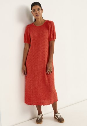 Frau mit dunklem Haar zu einem glatten Pferdeschwanz gebunden, trägt ein strukturiertes rotes Midikleid mit Puffärmeln und braunen Sneakers, steht vor einer schlichten weißen Wand.