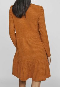 Femme aux cheveux noirs raides portant une robe orange-brun à manches longues, côtelée, avec une jupe froncée, vue de dos.