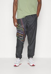 Man som bär svarta Nike-träningsbyxor med en färgglad konturerad logga på vänster ben, vita och röda sneakers och en ljusgrön t-shirt.