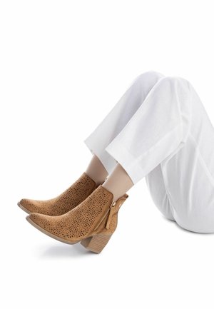 Stivaletti marroni traforati alla caviglia con tacco spesso e cerniera laterale, abbinati a pantaloni bianchi corti, che mettono in risalto texture e design.