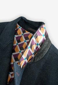 Mörkblå textiljacka med ett mönstrat foder i lila, gult och svart. Har en grå filtkrage och en strukturerad design.