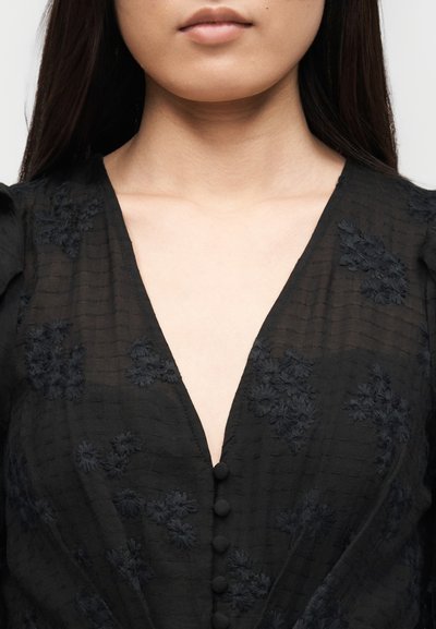 Γυναίκα με μαύρο μπλουζάκι σε σχήμα V με κεντήματα λουλουδιών και κουμπιά καλυμμένα με ύφασμα, που φαίνεται η κλείδα και το κάτω μέρος του προσώπου.