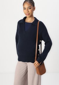 Navy knitted sweater with a V-neck, button accents on the side, paired with light ribbed pants and a brown crossbody bag.