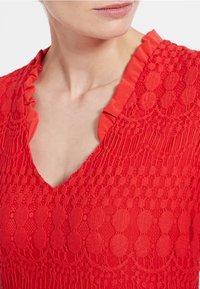 Červený pletený top s výstřihem do V a volánovým límcem, zdobený strukturovanými vzory kruhů a pruhů po celé látce.