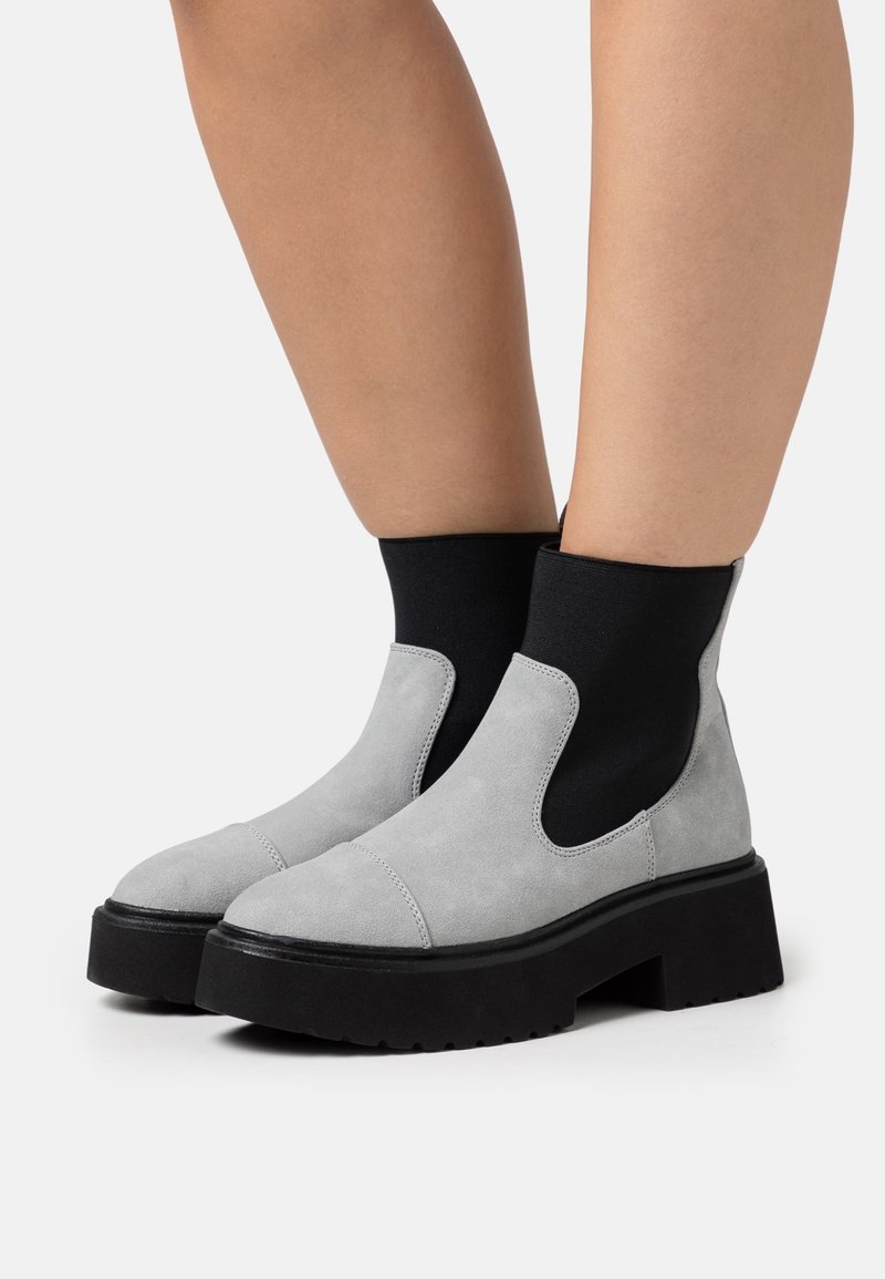 Harmaat mokkasaappaat, joissa on musta joustava sivupaneeli, paksu musta paksupohja ja pyöreä varvas. Tasainen pinta ja ompelu koristeena.