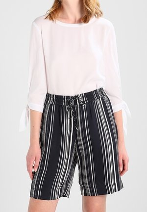 Blusa bianca con maniche a tre quarti e polsini legati, abbinata a pantaloni corti neri con righe bianche verticali e vita elastica.