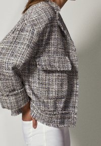 Strukturalna, tkana kurtka w odcieniach niebieskiego i szarego; z postrzępionymi brzegami i dużą kieszenią. Połączona z białymi spodniami.