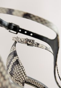 Chaussure à talons hauts avec un motif imprimé serpent en noir et crème, présentant un design élégant, une texture brillante et une bride ajustable à la cheville.