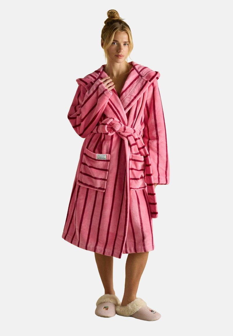 Różowy szlafrok w paski wykonany z miękkiego materiału, z kapturem, wiązaniem w talii i dwoma przednimi kieszeniami. Noszony z jasnymi, puszystymi klapkami.