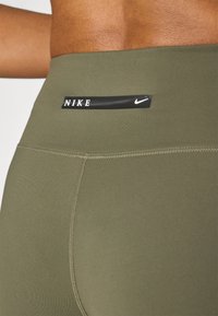 Tummanvihreät urheiluleggingsit, joissa on sileä pinta, musta Nike-logo vyötäröllä ja näkyvät saumat.