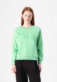 Zeleno sweatshirt z embosiranim napisom "PARIS" spredaj, okroglim izrezom, z ribanim zaključkom rokavov in roba. Nosi se preko črnih hlač.