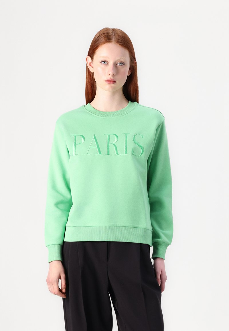Zeleno sweatshirt z embosiranim napisom "PARIS" spredaj, okroglim izrezom, z ribanim zaključkom rokavov in roba. Nosi se preko črnih hlač.