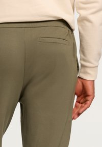 Olive green pants with a smooth texture and elastic waistband, featuring a single welt pocket on the back and a relaxed fit.